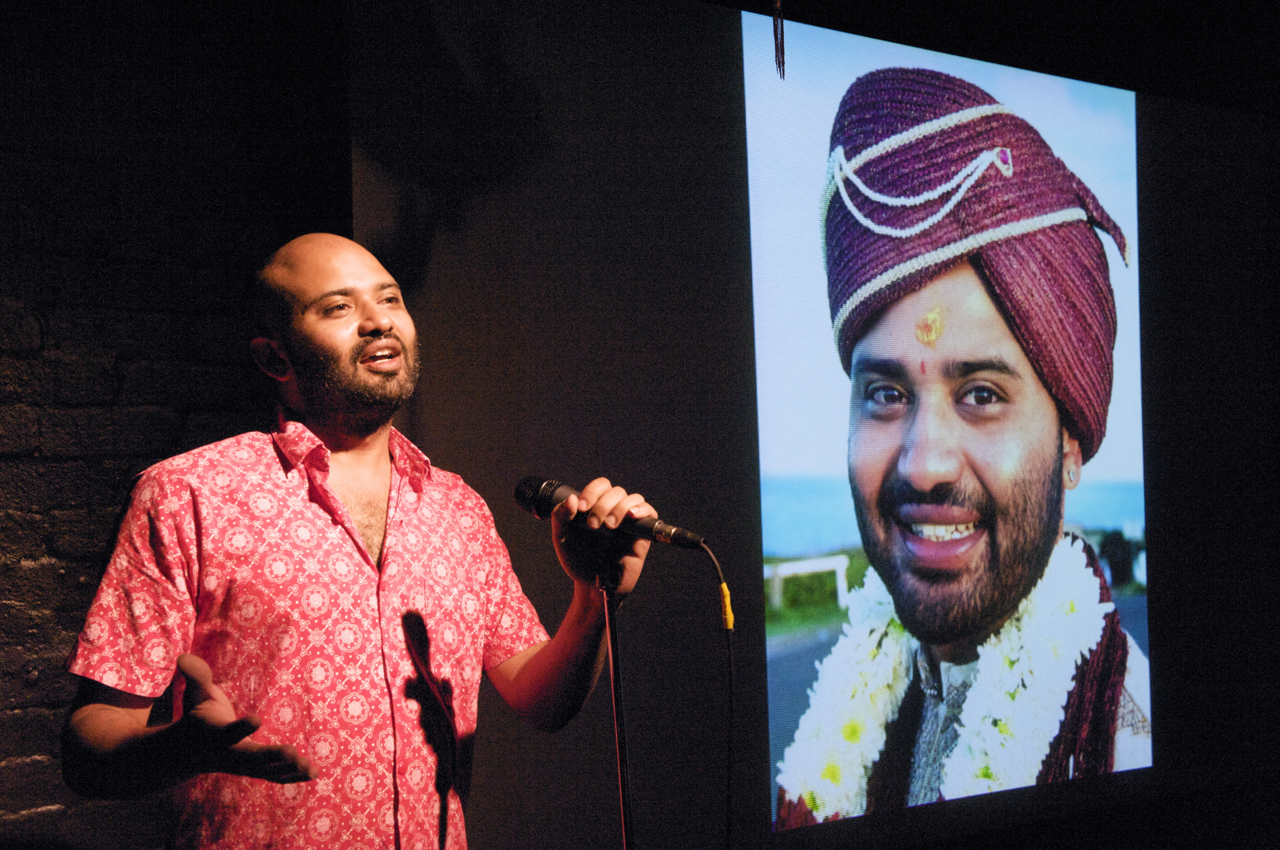 Sunil in red shirt at microphone in front of a picture of himself in a turban